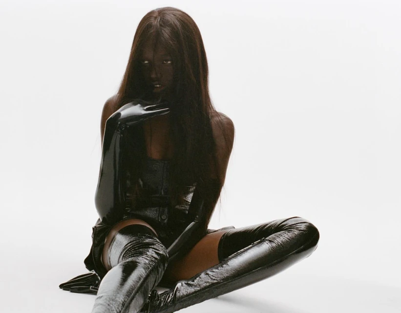 Woman in a glossy black outfit, including a corset, gloves, and thigh-high boots, poses in a studio against a stark white backdrop.