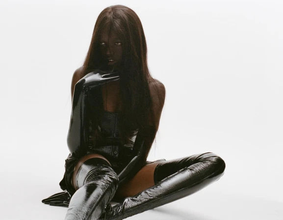 Woman in a glossy black outfit, including a corset, gloves, and thigh-high boots, poses in a studio against a stark white backdrop.