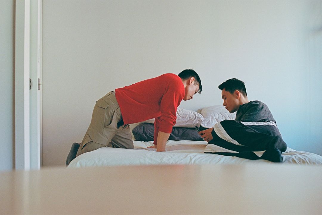Two Asian men are on a bed in a brightly lit indoor setting. One man is kneeling, and the other is sitting and looking at a mobile phone.