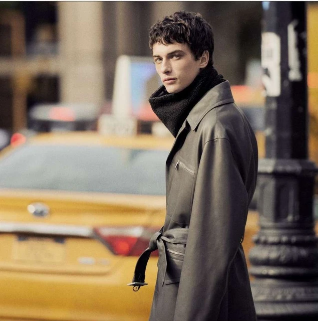 A young adult man in a long grey trench coat and black scarf stands on a city street with a yellow taxi cab in the background.