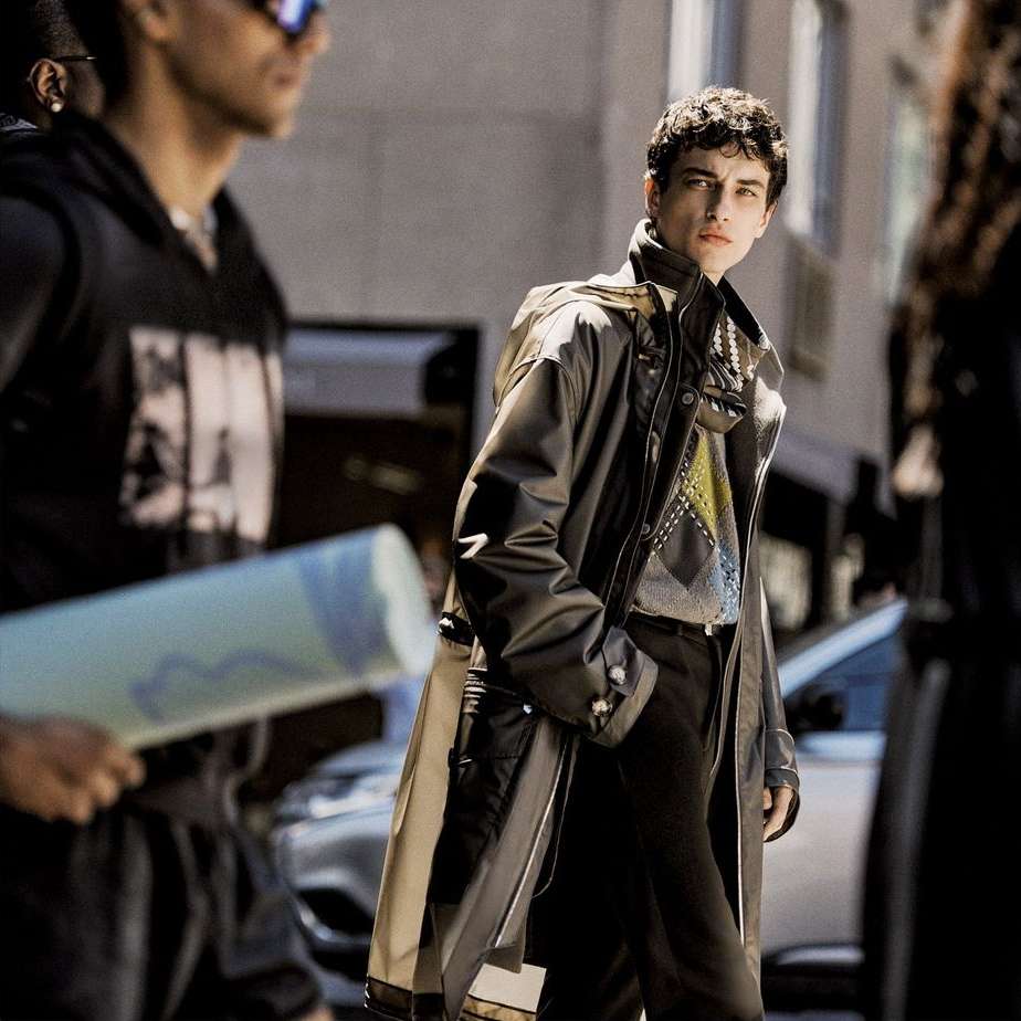 A young adult man walks on a city street wearing a long, stylish black coat and patterned sweater, flanked by blurred figures.