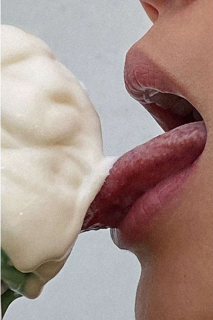 Close-up of a person with their tongue extended, licking white foam or cream from a green object against a light grey background.