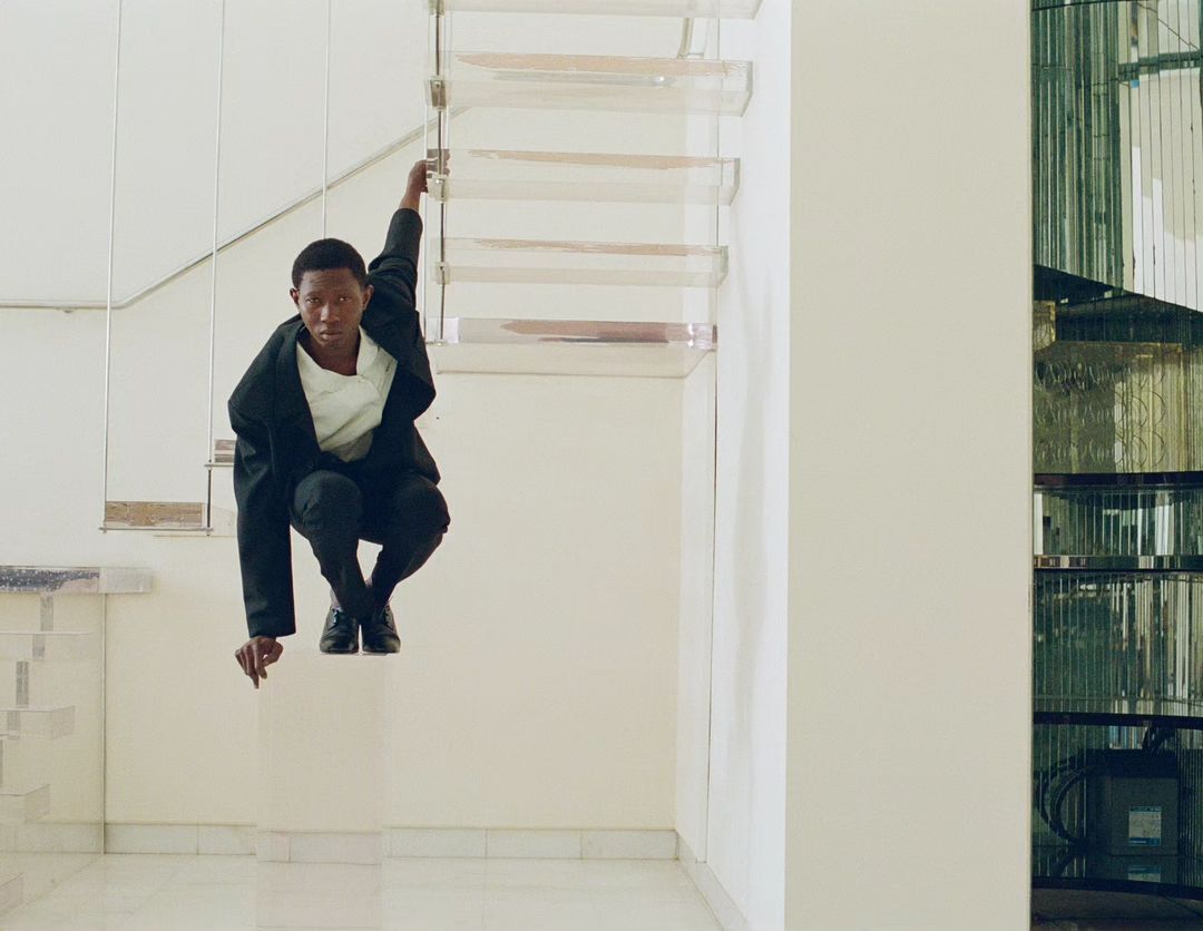 A young man in a black suit jacket and black pants squats in an indoor space with a mirrored wall and a light-colored floor and walls.