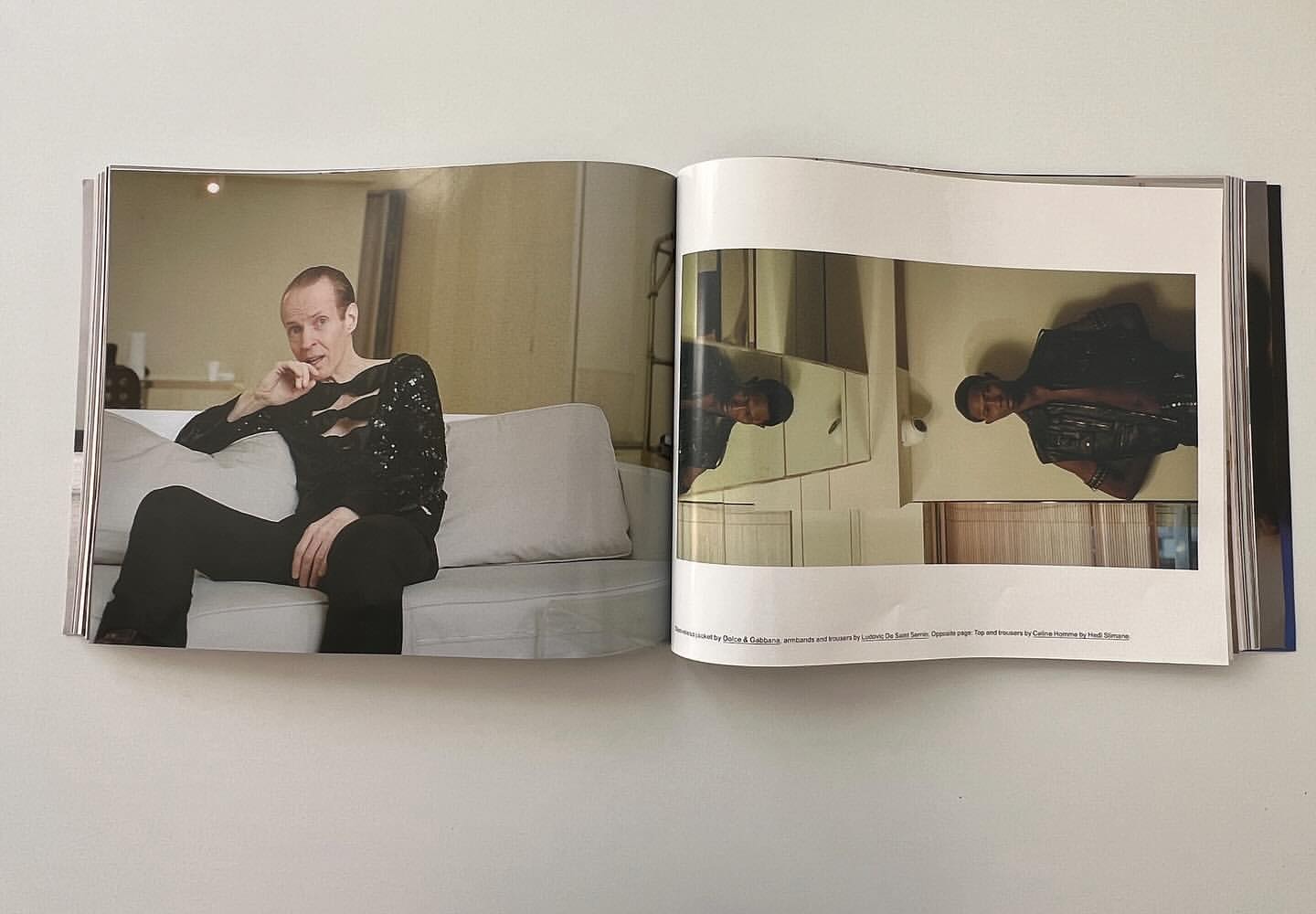 Fashion editorial image features a man with short hair wearing a black sparkly cut-out shirt and trousers, sitting on a white sofa.
