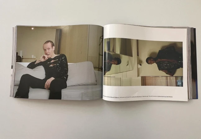 Fashion editorial image features a man with short hair wearing a black sparkly cut-out shirt and trousers, sitting on a white sofa.