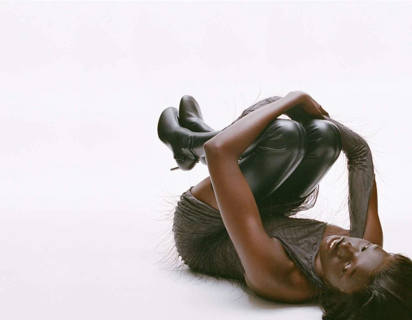 Woman with dark skin and hair poses with legs up wearing black boots and a gray top and leggings on a white background.
