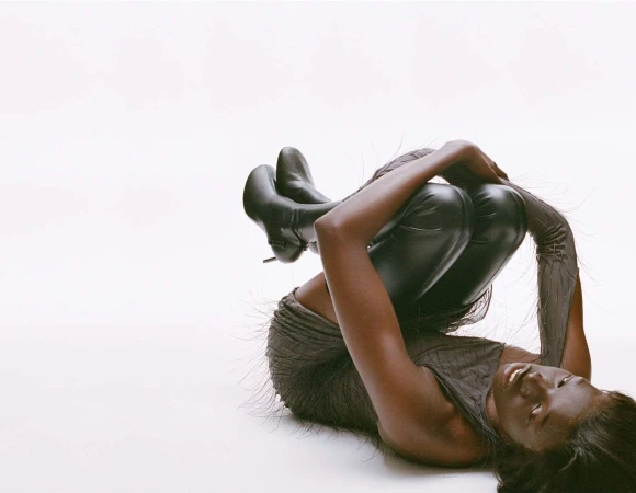 Woman with dark skin and hair poses with legs up wearing black boots and a gray top and leggings on a white background.