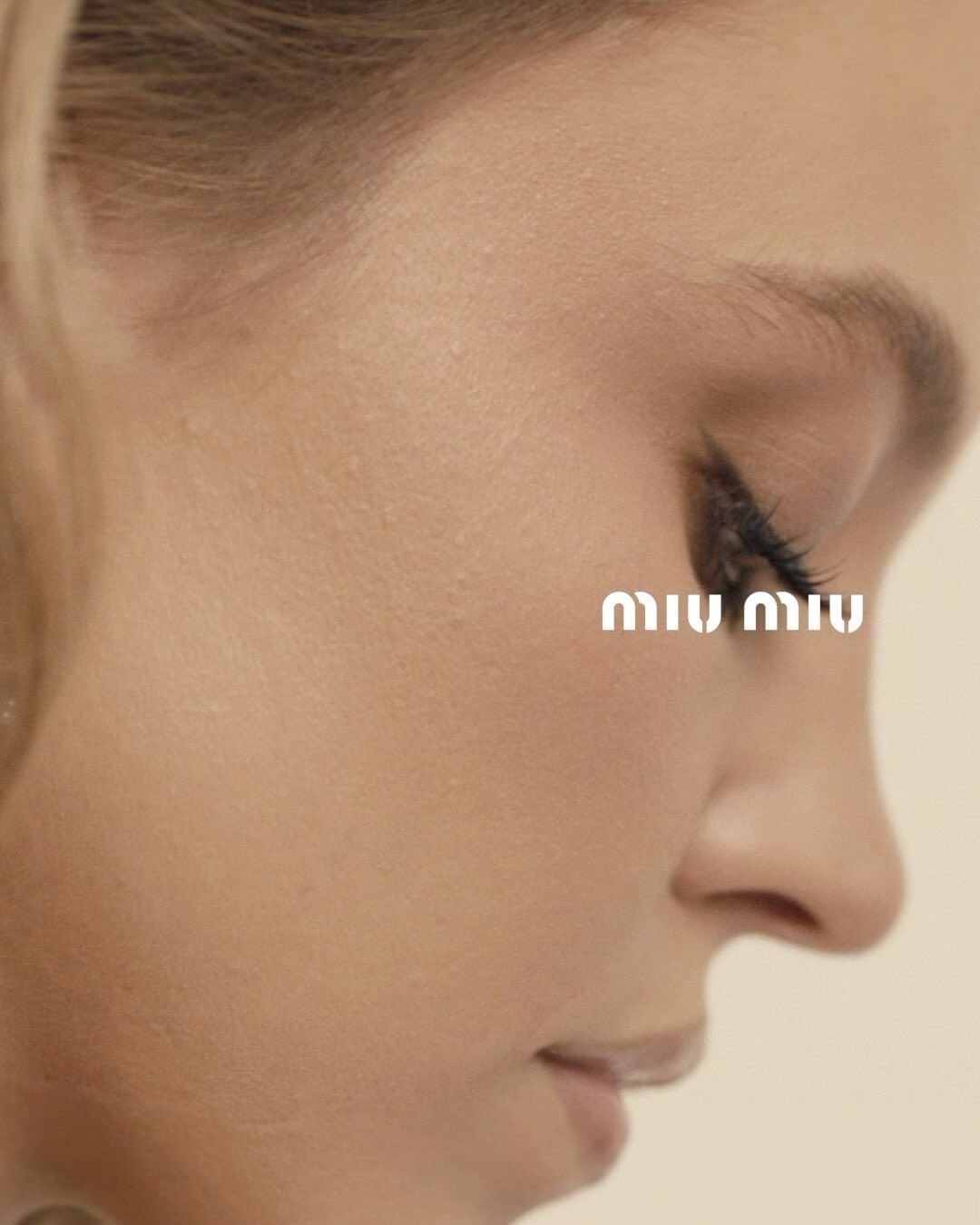 Close-up shot of a blonde adult woman's face in a beauty editorial ad for Miu Miu, showcasing makeup and skincare in a photo studio setting.