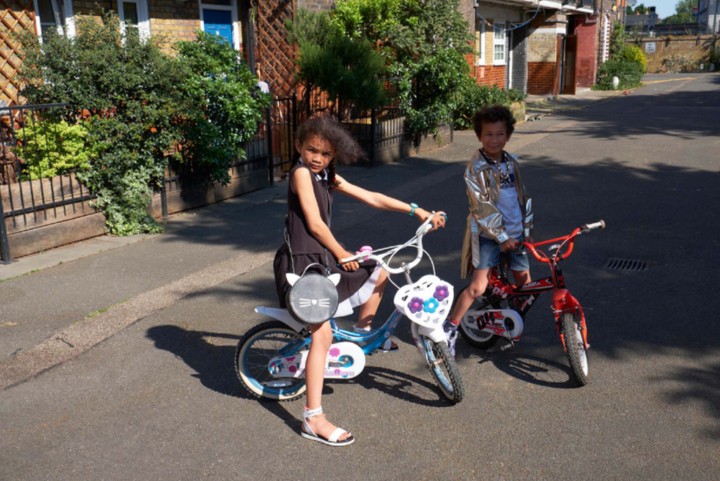 Two kids pose on their bikes in an urban environment on a sunny day. A girl in black sits on a blue and white bike and a boy in a shiny jacket.