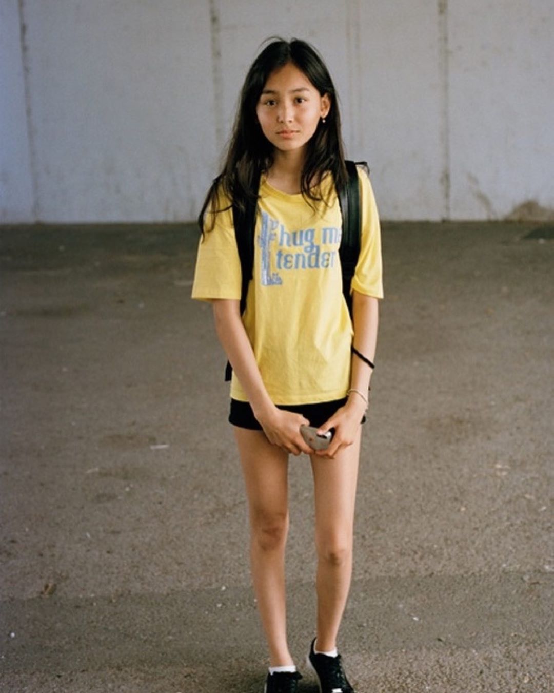 Portrait of an Asian woman in a yellow t-shirt with dark hair, standing in an urban setting holding a cell phone.