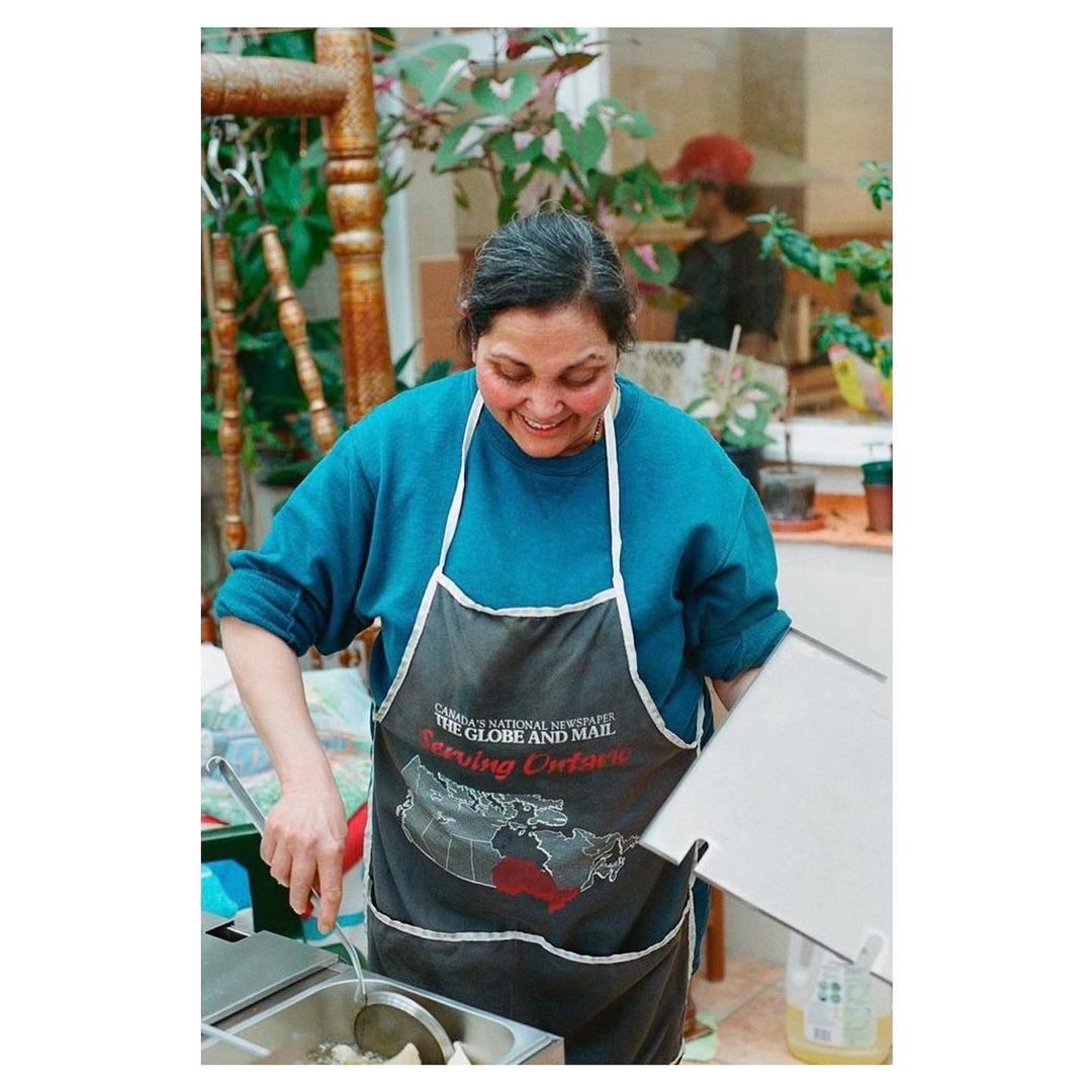 Indoor shot of a woman wearing an apron, standing near a food preparation area, possibly cooking or serving food, with plants visible.