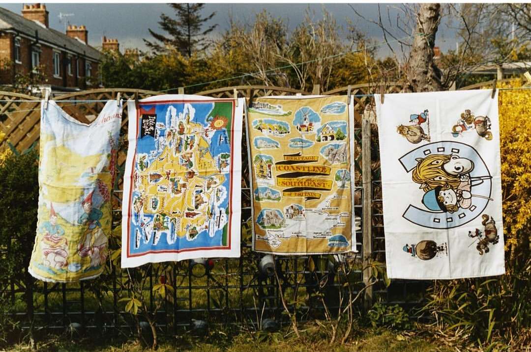 Outdoor shot of colorful souvenir tea towels hanging on a clothesline, depicting cartoon characters and maps, against a backdrop of houses and trees.