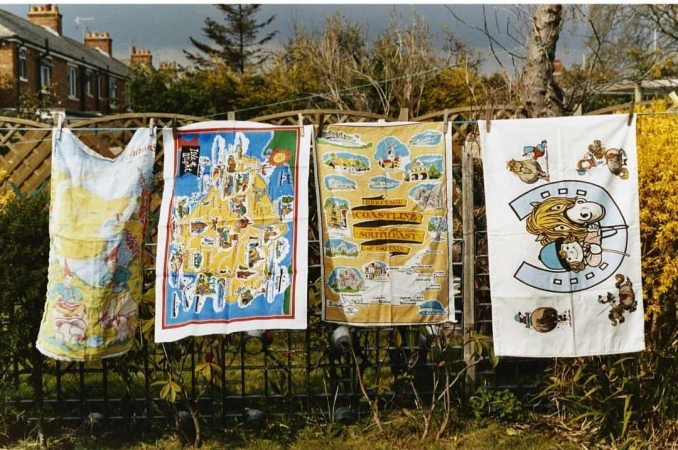 Outdoor shot of colorful souvenir tea towels hanging on a clothesline, depicting cartoon characters and maps, against a backdrop of houses and trees.