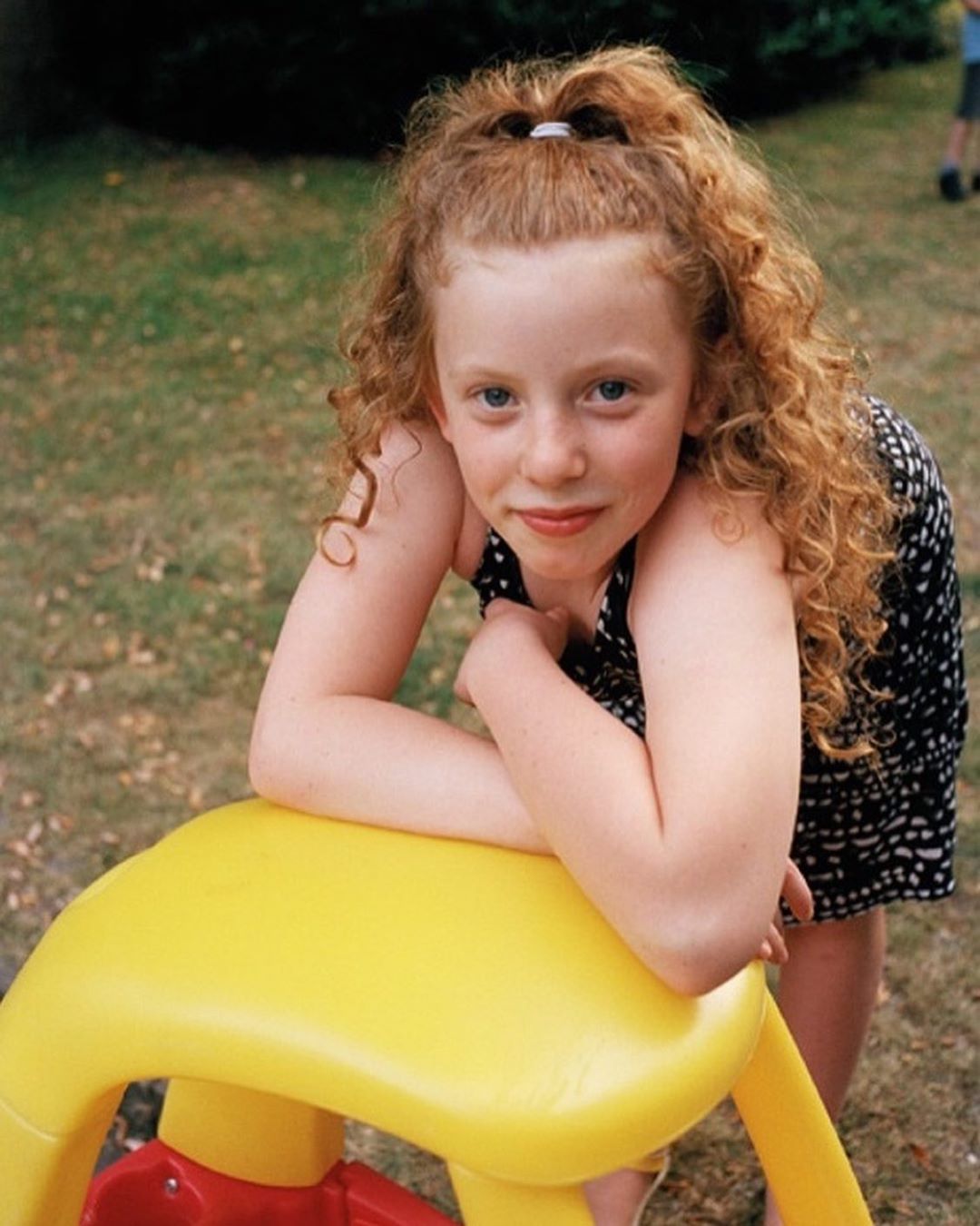 Portrait of a red-haired girl wearing a black and white patterned dress, leaning on a yellow and red play structure outdoors.