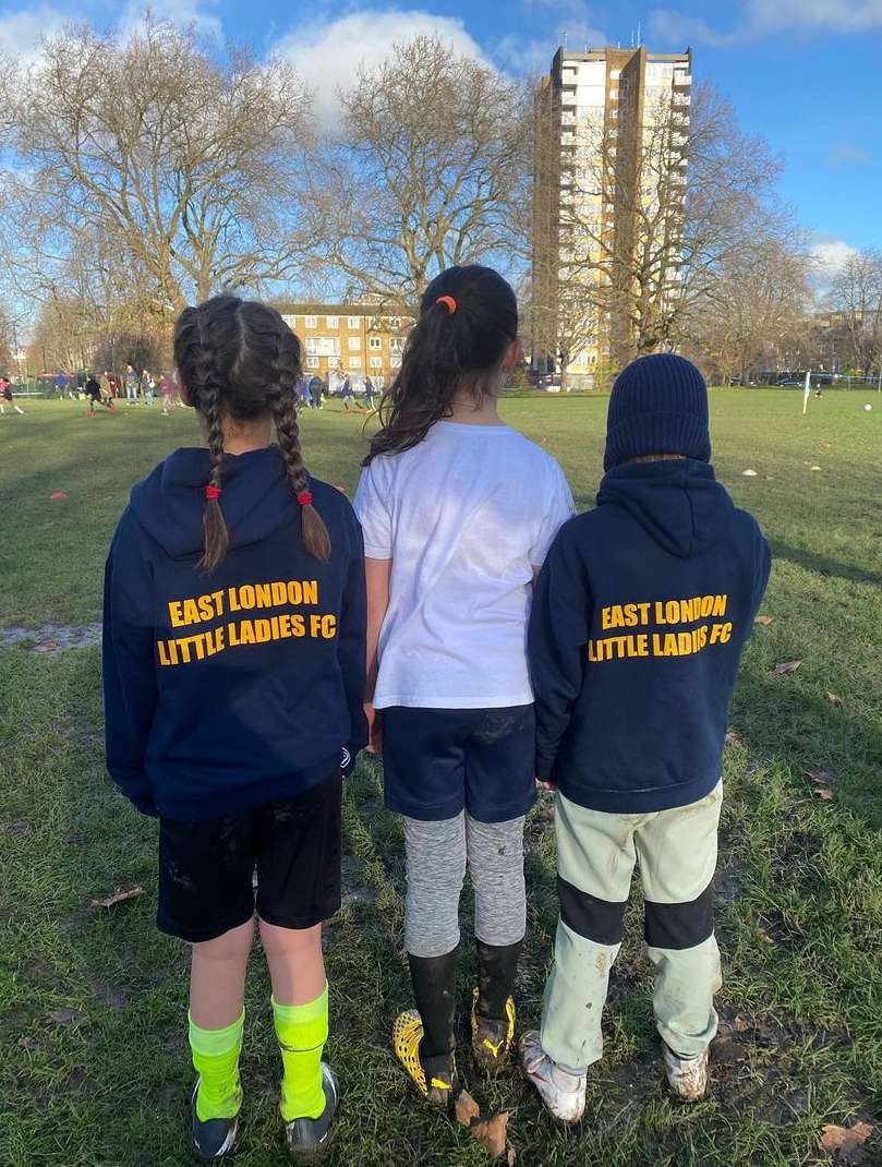 Three young soccer players in matching hoodies stand on a grass field in daylight, with city buildings and bare trees in the background.