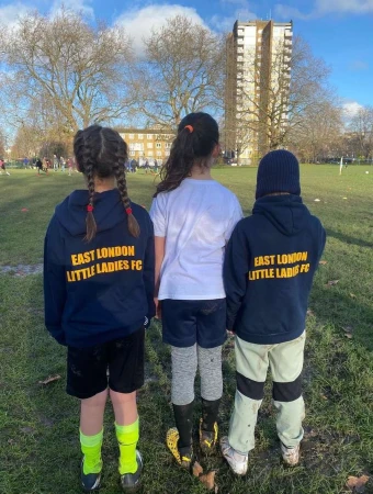 Three young soccer players in matching hoodies stand on a grass field in daylight, with city buildings and bare trees in the background.