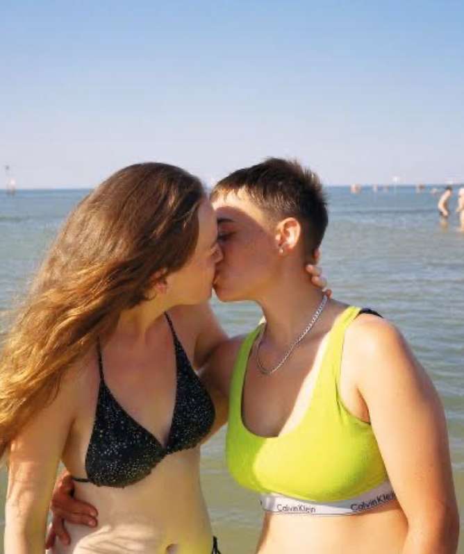 Two young adult women in bikinis embrace and kiss on a sunny beach with other people in the background swimming in the ocean.