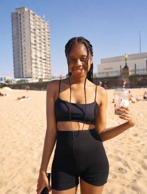 Woman in black athletic wear poses on beach with drink, bright sunlight, high-rise building in background, medium shot, outdoor daylight.