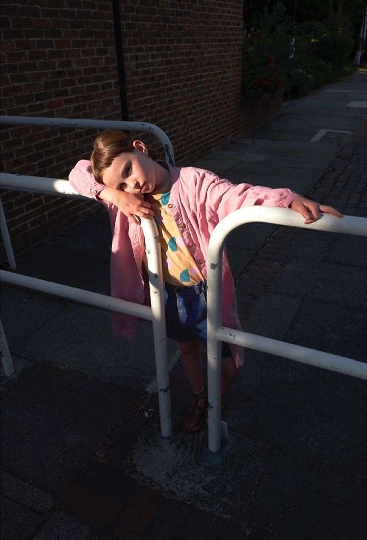 A young child in a pink jacket rests against a white railing next to a brick wall outdoors, with sunlight and shadows highlighting the scene.