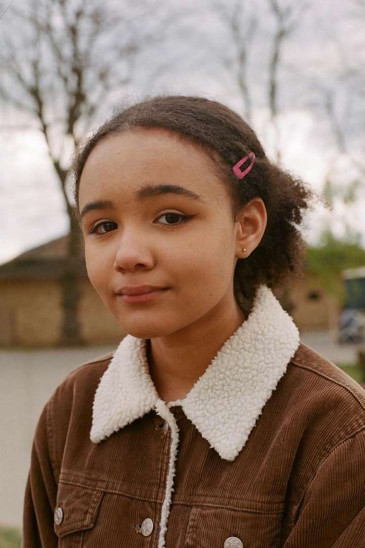 Portrait of a young woman with curly hair wearing a brown corduroy jacket with a sherpa collar, standing outside on an overcast day.