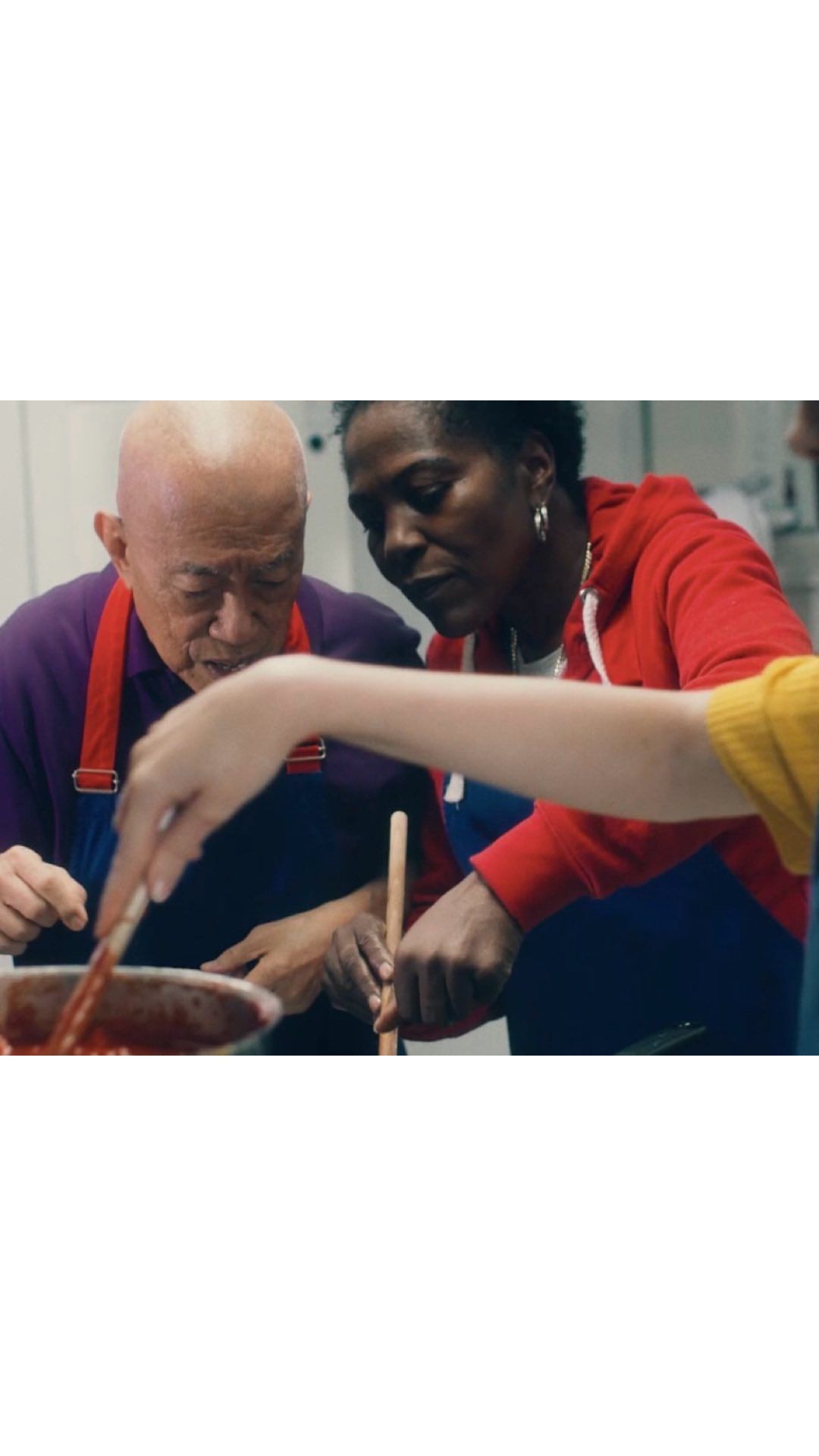 Two adults and a child stirring food in a pot indoors, wearing aprons and casual clothes, under bright lighting, with a shallow depth of field.