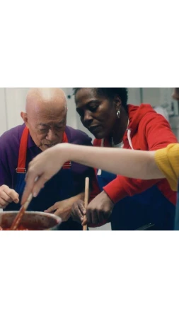 Two adults and a child stirring food in a pot indoors, wearing aprons and casual clothes, under bright lighting, with a shallow depth of field.