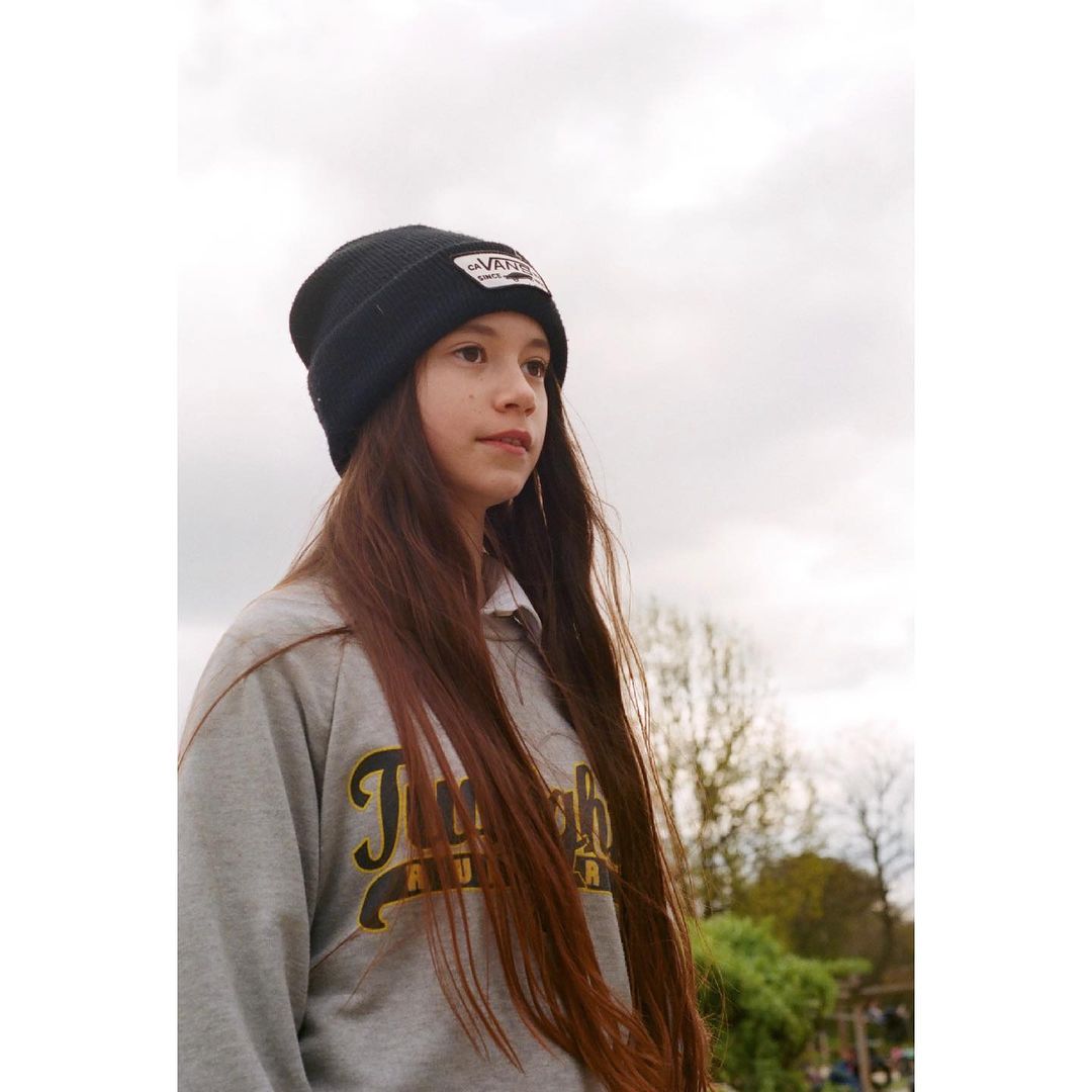 A young woman stands outside wearing a black beanie and gray sweatshirt, with long brown hair and a serene expression against a cloudy sky.