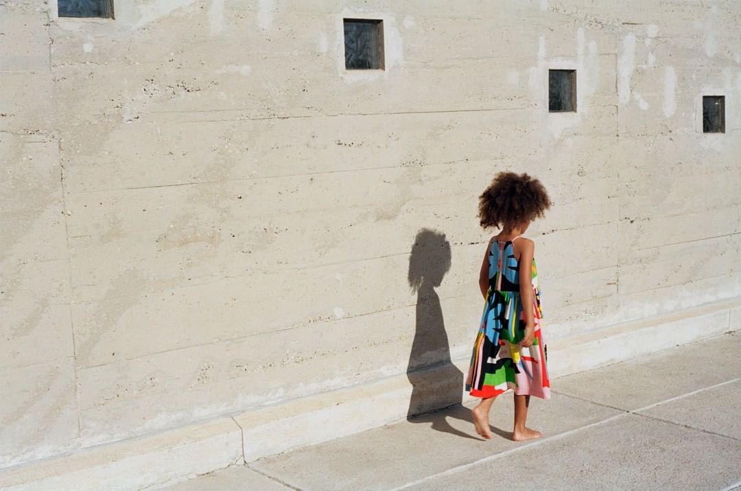 Back view of a young girl with afro wearing colorful dress walking barefoot along sidewalk in front of textured concrete wall with windows.