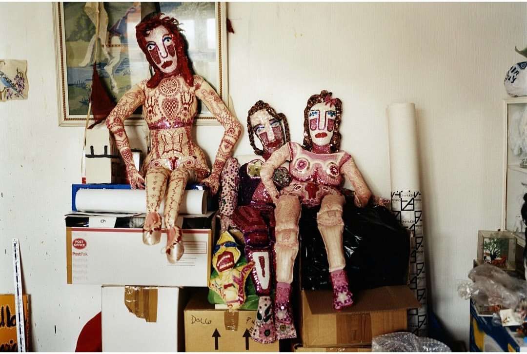Three handcrafted art dolls sit on stacked cardboard boxes in an indoor setting, exhibiting a mix of artistic and whimsical design elements.