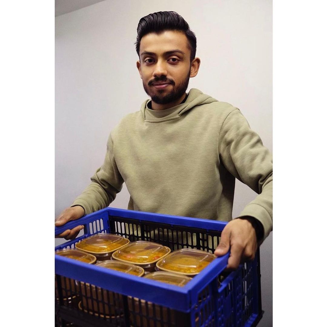 A man holds a blue crate full of food containers in an indoor environment, showcasing food delivery or preparation with a neutral backdrop.
