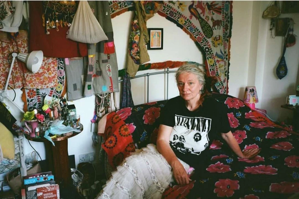 Adult woman with silver hair in a black Nirvana shirt sits on a bed with a red floral pattern in a bedroom full of textiles.