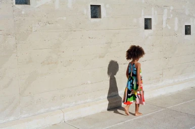 Young child in a colorful patterned dress walking barefoot along a concrete wall with small square windows.