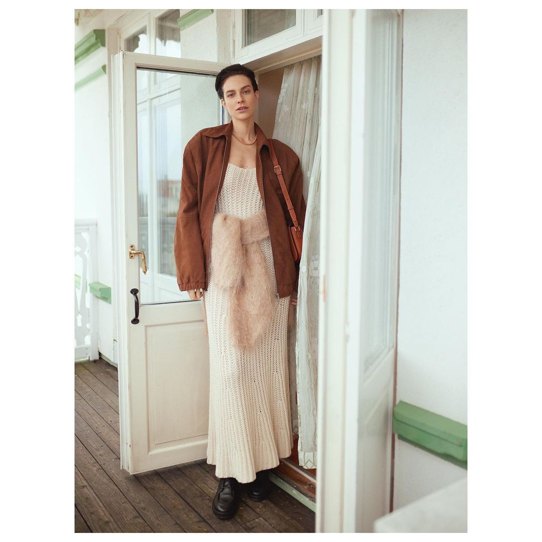 An adult woman poses in a knitted dress and brown jacket standing in a doorway, with a light, airy aesthetic and soft, neutral color palette.