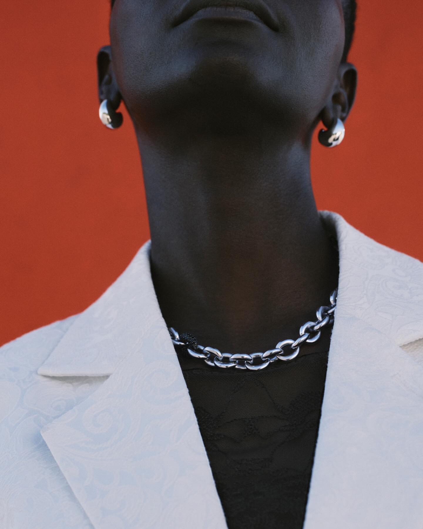 Close-up of an adult with dark skin wearing silver chain necklace and earrings with a white blazer against an orange background.