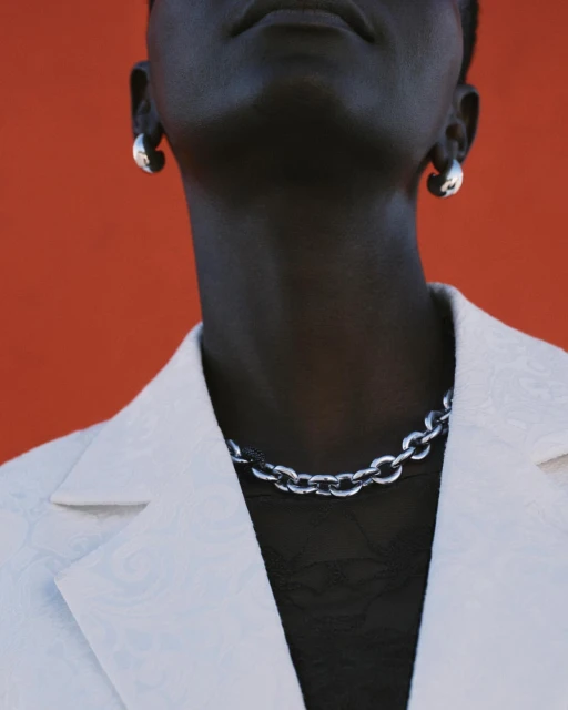 Close-up of an adult with dark skin wearing silver chain necklace and earrings with a white blazer against an orange background.