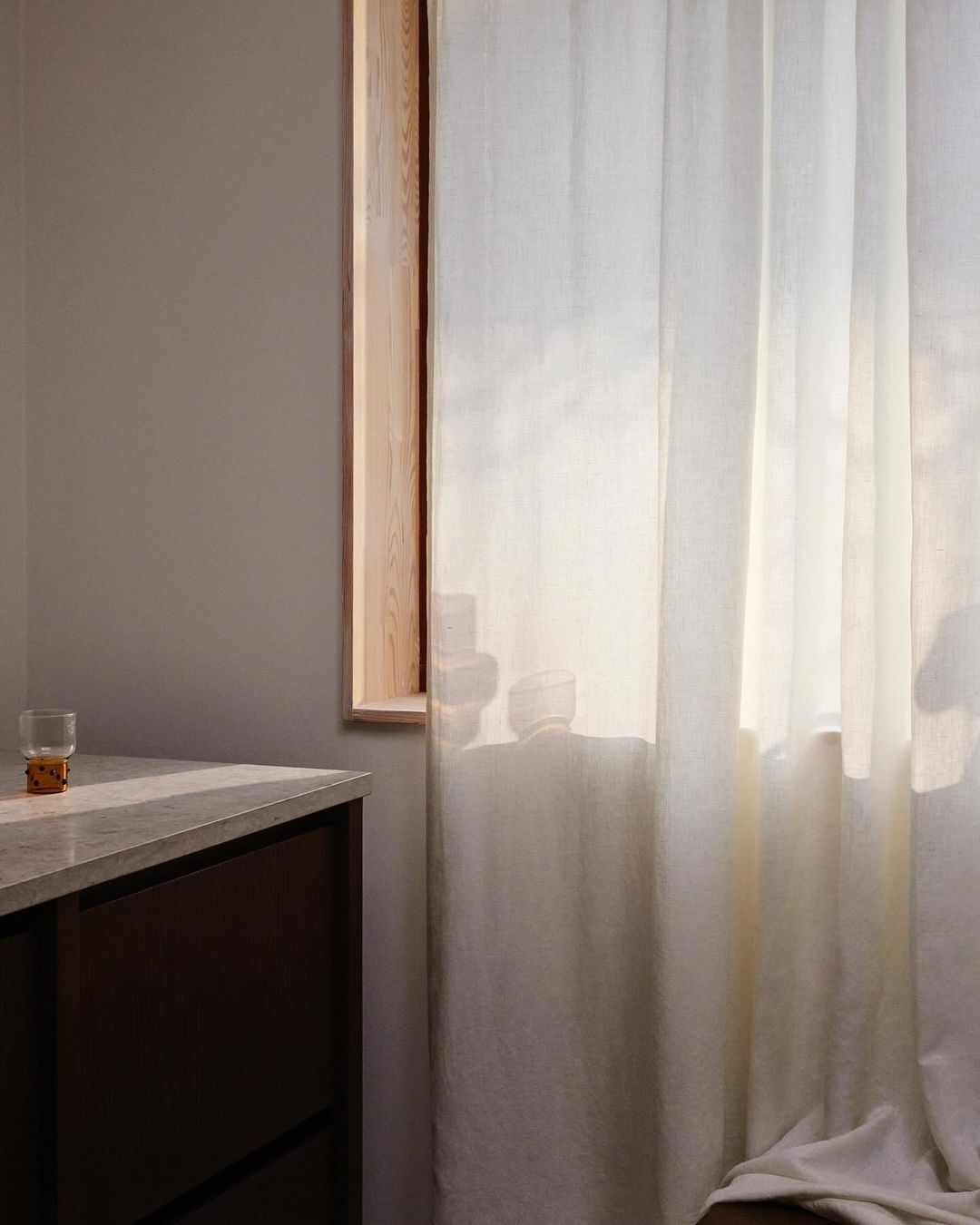 Serene interior with natural light shining through sheer white curtains, a wooden window frame, grey walls, and a modern dark cabinet.