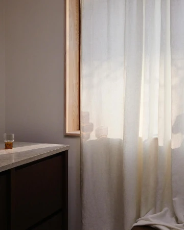 Serene interior with natural light shining through sheer white curtains, a wooden window frame, grey walls, and a modern dark cabinet.