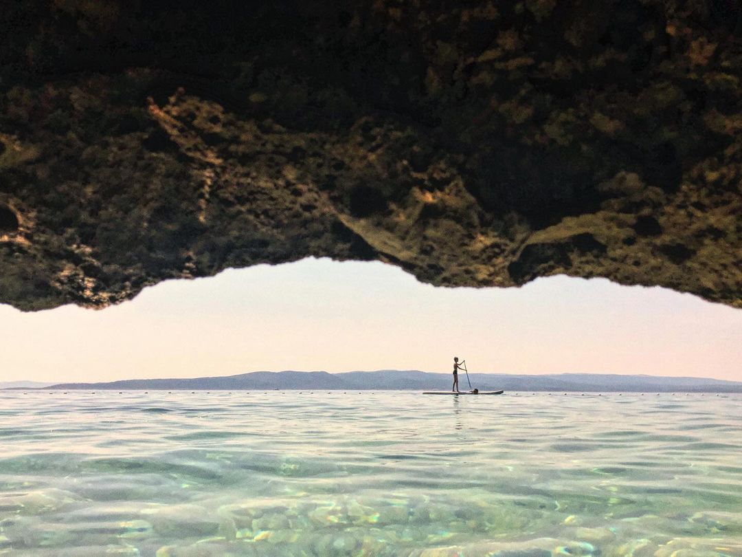Person paddleboarding on calm water with a distant coastline, seen from the perspective of an underwater cave opening.
