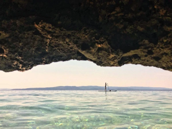 Person paddleboarding on calm water with a distant coastline, seen from the perspective of an underwater cave opening.