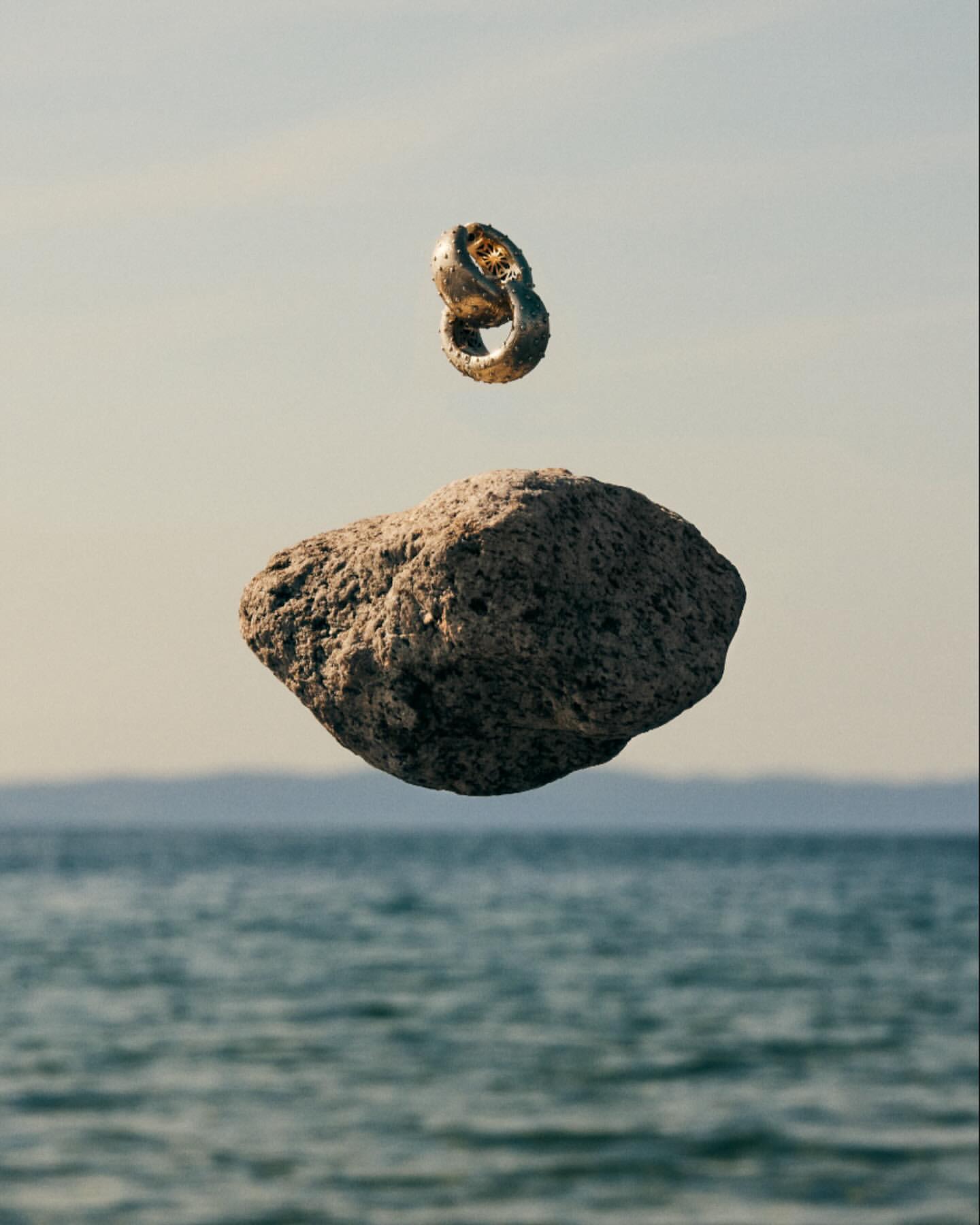 Art photograph of a gold ring hovering above a large rock, with a light blue sky and rippling water in the background.