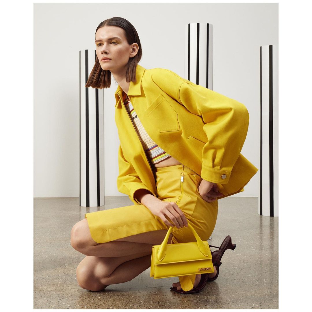 A woman in a yellow outfit kneels and holds a matching purse, with vertical black and white striped pillars in the background.