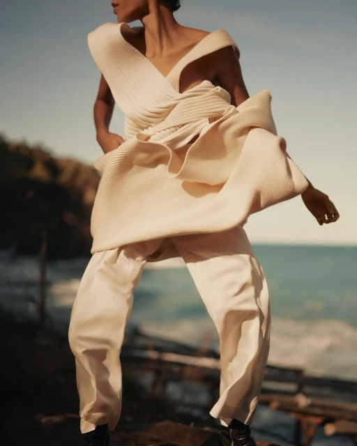 Fashion photography showcases a stylish adult female in an oversized knit top and trousers on a rocky coast with a pale blue sea.