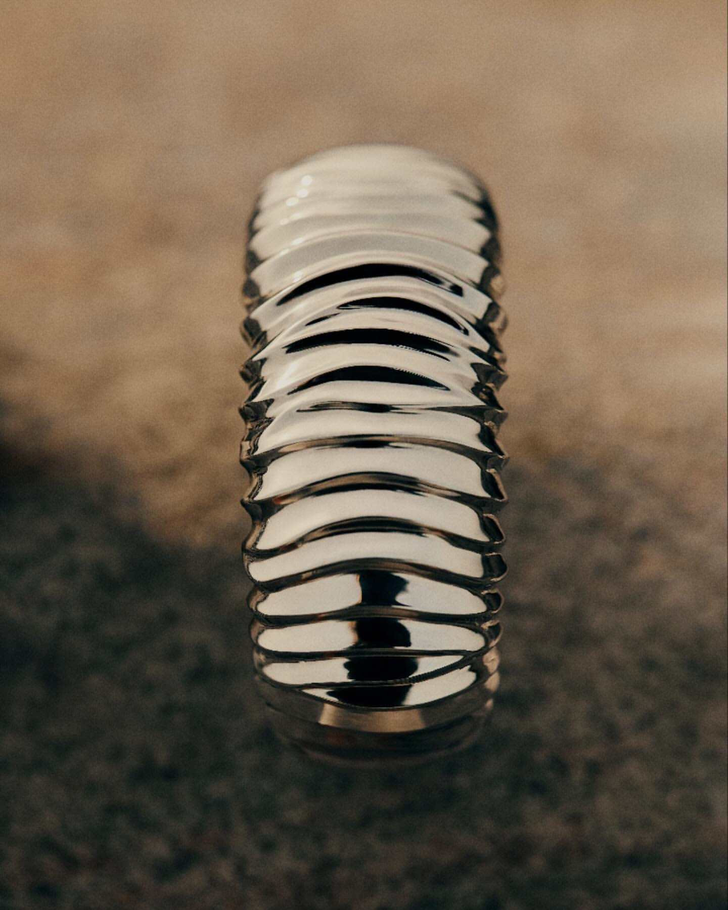 Close-up of a ridged, metallic silver object with a blurry, mottled brown and beige background, showing texture and abstract form.