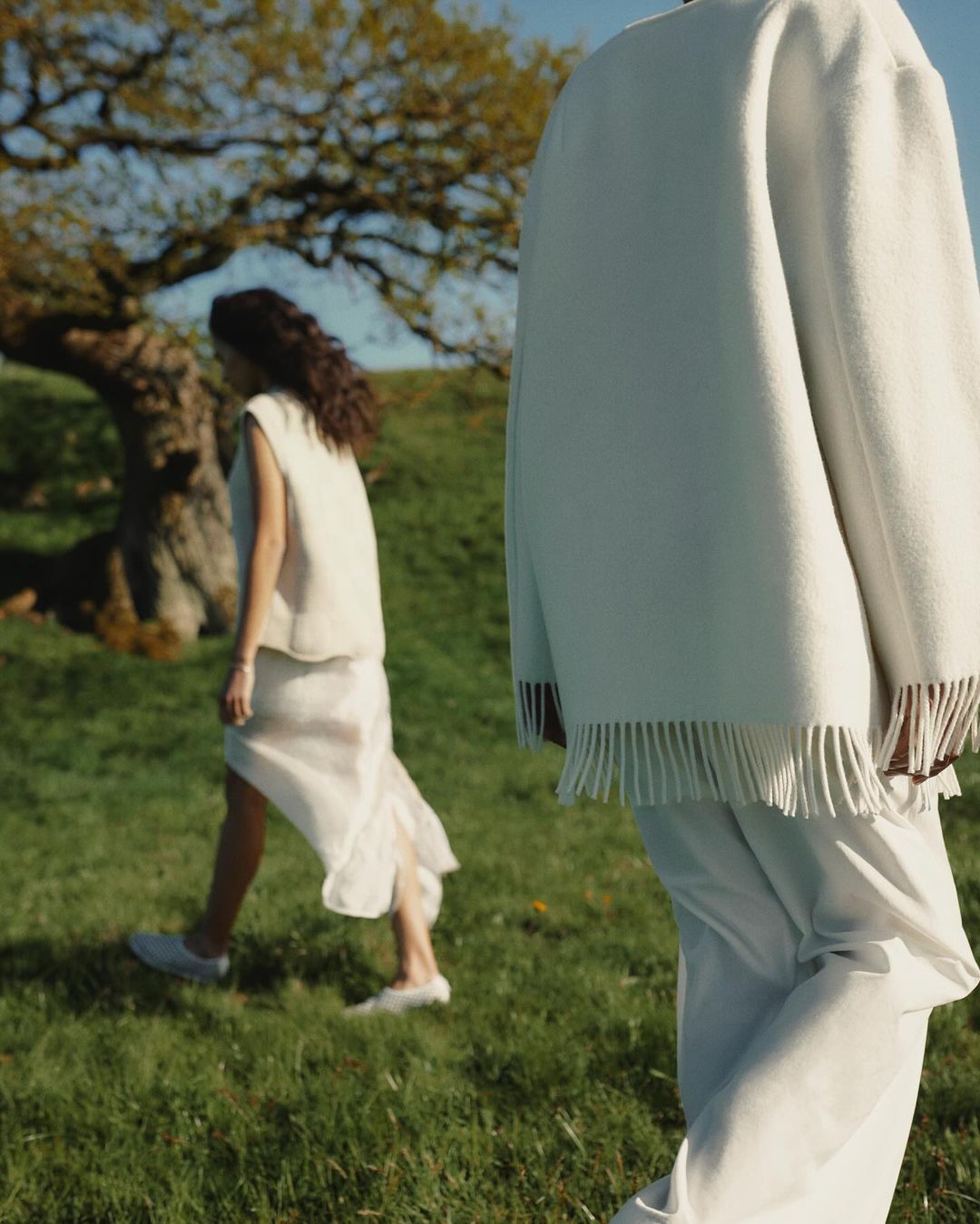 Two women in a meadow, dressed in light-colored sleeveless tops and skirts; one is in focus, the other blurry, near a tree and blue sky.