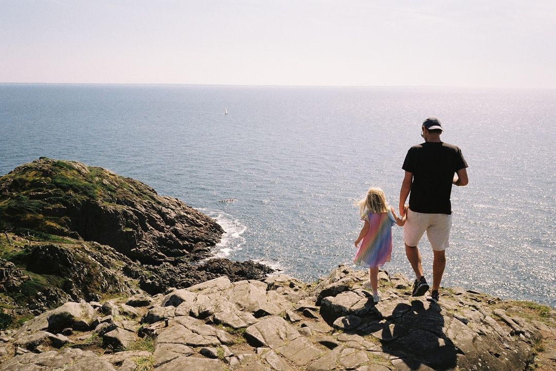 Man and child holding hands, walking on a rocky cliffside overlooking the ocean under a sunny sky, with a sailboat visible in the distance.
