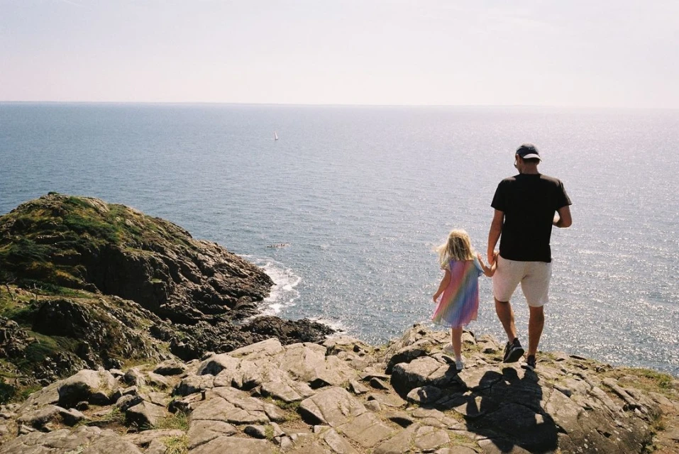 Man and child holding hands, walking on a rocky cliffside overlooking the ocean under a sunny sky, with a sailboat visible in the distance.