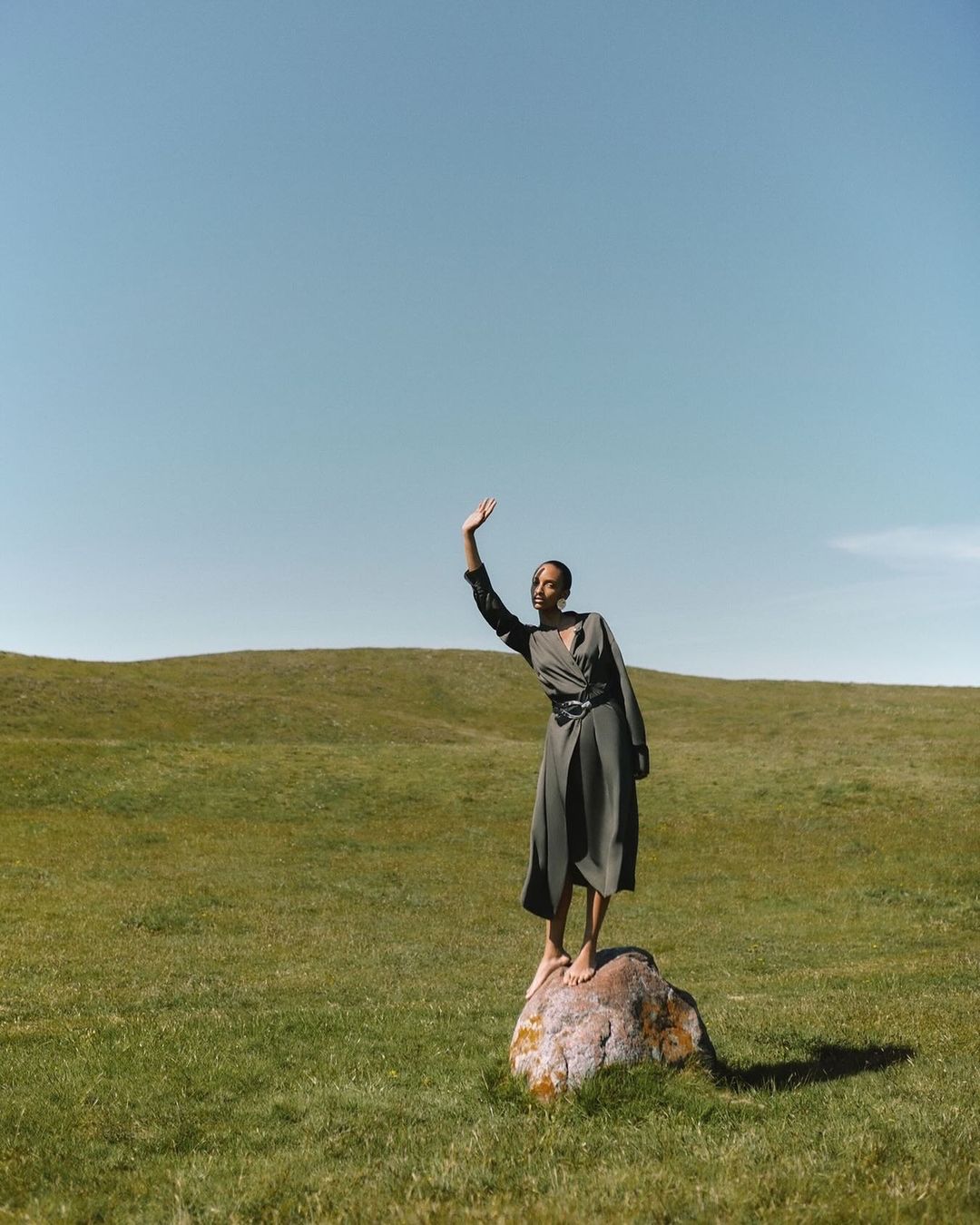 Adult woman in olive dress stands barefoot on a boulder in a green meadow with blue sky, waving in a natural light fashion shot.