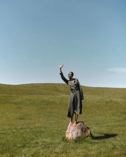 Adult woman in olive dress stands barefoot on a boulder in a green meadow with blue sky, waving in a natural light fashion shot.