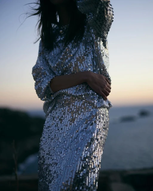 Woman in a shimmering silver sequin dress stands outdoors at dusk near the ocean, modelling fashion in a lifestyle setting.