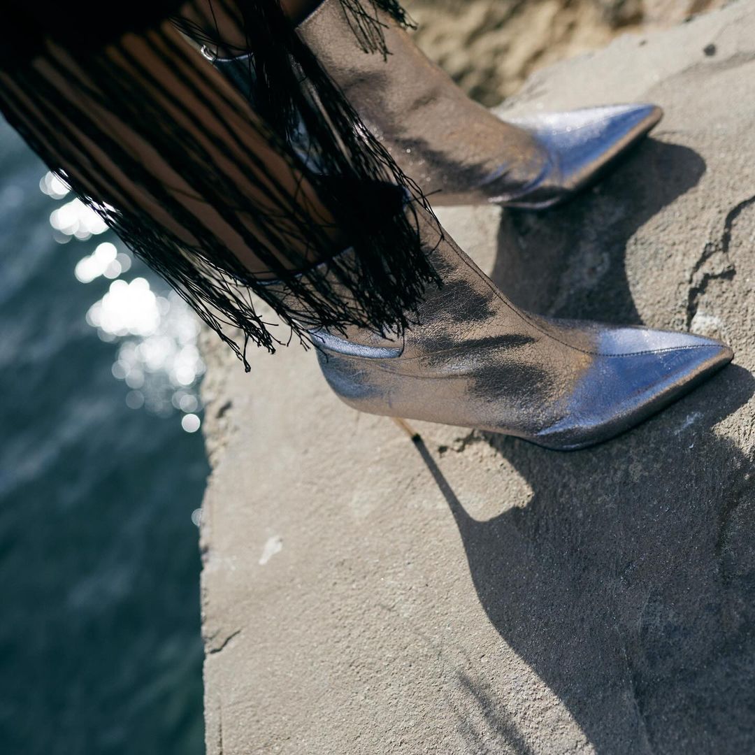 Silver ankle boots with stiletto heels and fringe detail on a concrete surface with water in the background create a fashion-forward look.